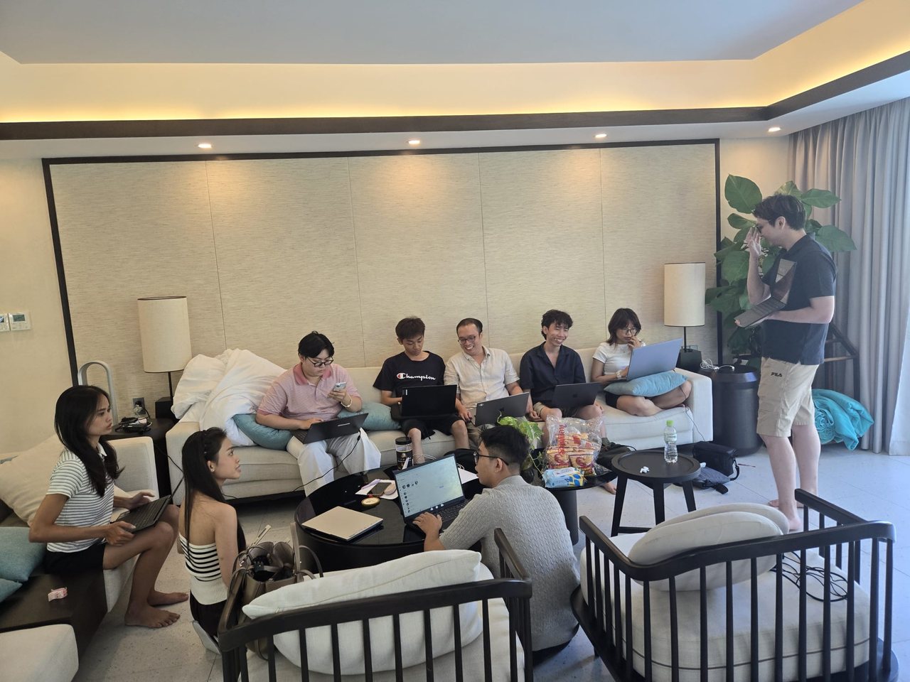 Accountants and developers working on laptops around a living room during the team's hackathon in Vietnam, with one team member standing to present.